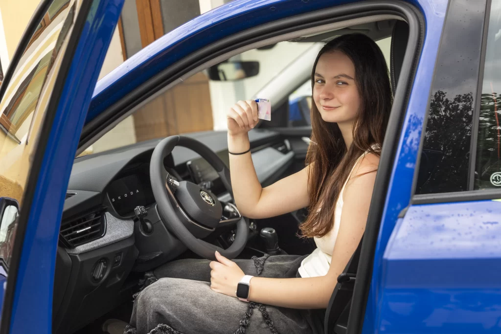 studentka autoškoly sedí ve voze a ukazuje čerstvě získaný řidičský průkaz